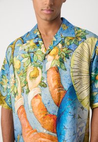 Camisa de manga corta y botones con un colorido patrón artístico de manos y frutas sobre un fondo azul. Material de algodón.