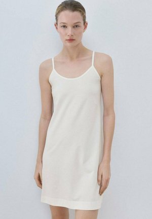 Mujer joven de piel clara y cabello recogido vestida con un vestido blanco sin mangas, con un fondo liso gris claro.