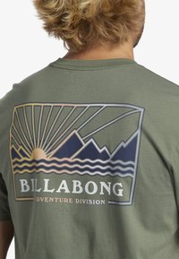 Olive green cotton t-shirt featuring a rear graphic with a sun, mountains, and waves in shades of navy and gold. Includes "BILLA BONG ADVENTURE DIVISION."