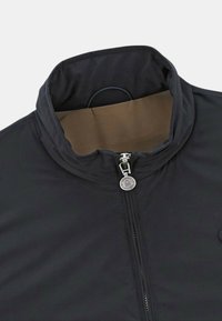 Veste noire zippée avec un col haut et une texture lisse. Dotée d'une tirette de fermeture éclair circulaire en argent et d'une doublure intérieure marron.