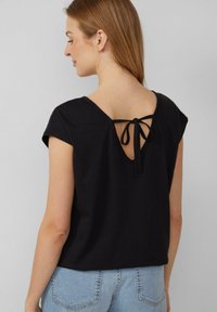 Vrouw met haar rug naar de camera, gekleed in een zwarte korte mouw top met een lage rug en een strikdetail, en lichte spijkerjeans.