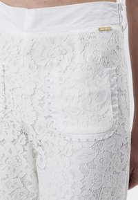 Witte kanten broek met een bloemmotief, een effen witte tailleband en een zak versierd met kleine parelaccenten.