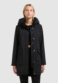 Jeune femme portant un long manteau noir à capuche avec boutons-pression et poches, debout devant un fond uni gris clair.