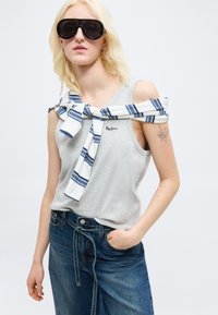 Ξανθιά γυναίκα που φοράει μαύρα μεγάλα γυαλιά ηλίου, γκρι αμάνικη μπλούζα, ριγέ πουλόβερ δεμένο στους ώμους και μπλε τζιν με το ένα χέρι στην τσέπη.