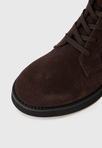 Pruunist nahast lace-up saapad musta kummist taldaga, valge tausta ees, näidates lähedalt varba ja paelte ala.