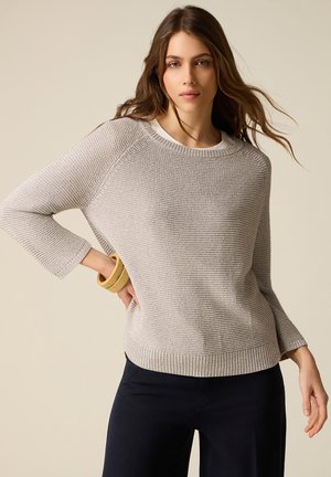 Kvinde med langt brunt hår iført en lys beige tekstureret striktrøje, sorte bukser og træ-armbånd, stående foran en ensfarvet baggrund.