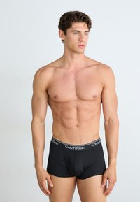 Zwarte Calvin Klein boxershorts met een strakke pasvorm, voorzien van een gladde textuur en een gebrandmerkte tailleband. Model staat tegen een lichte achtergrond.