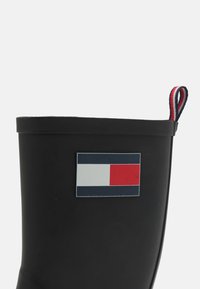 Botte en caoutchouc noire avec une surface lisse, présentant un logo rectangulaire en bleu, blanc et rouge. Comprend une sangle à rayures à l'arrière.