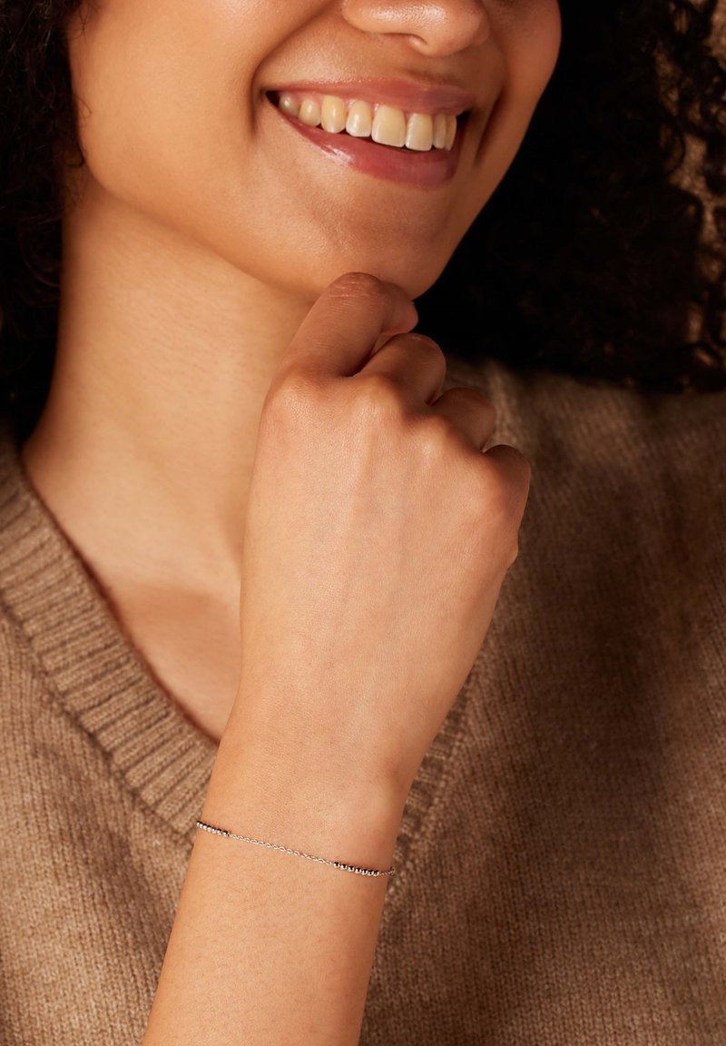Zilveren kettingarmband met kleine ronde kralen, gedragen om een pols. De achtergrond toont een zachte bruine trui en een gedeeltelijke glimlach.