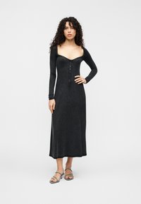 Μαύρο μακρυμάνικο φόρεμα από ριμπ ύφασμα, με τετράγωνη λαιμόκοψη και κούμπωμα κουμπιών μπροστά, με ρέουσα σιλουέτα μέχρι τους αστραγάλους.