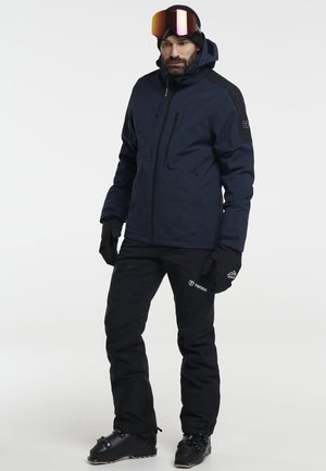 Man in een marineblauwe jas, zwarte skibroek, zwarte handschoenen, helm en skischoenen, staand tegen een effen witte achtergrond.