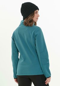Teal softshell jack met een hoge kraag en een aansluitend ontwerp. Heeft lange mouwen en subtiele stikdetails. Gekozen met een zwarte gebreide beanie.