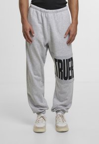 Sivé bavlnené tepláky s elastickým pásom a zúženými členkovými manžetami majú veľký čierny grafický nápis "TRUER" na dolnej ľavej nohe.