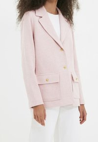 Blazer texturé rose avec un col à revers, design double boutonnage, deux poches frontales et accents de boutons dorés.