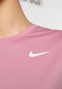 Růžové sportovní tričko s texturovanou tkaninou, se zaobleným výstřihem a malým bílým logem Nike ve tvaru swoosh na levé části hrudníku.