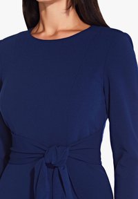 Sotiharma sininen mekko pitkillä hihoilla, pyöreällä kaula-aukolla ja sidottavalla vyökoristeella. Valmistettu sileästä kankaasta, jossa on räätälöity istuvuus.