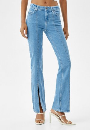 Widok z przodu kobiety noszącej jasnoniebieskie dżinsy z wysokimi rozcięciami przy przednich krawędziach oraz białe sandały na wysokim obcasie z paskami.