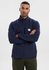 Chaqueta de forro polar azul marino con cuello alto, cierre frontal completo y bolsillos laterales. Presenta una pequeña etiqueta de la marca en el pecho. Textura suave.