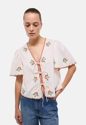 Femme aux cheveux attachés porte un chemisier blanc avec des broderies florales rouges et vertes et des fermetures à nouer sur un jean bleu.