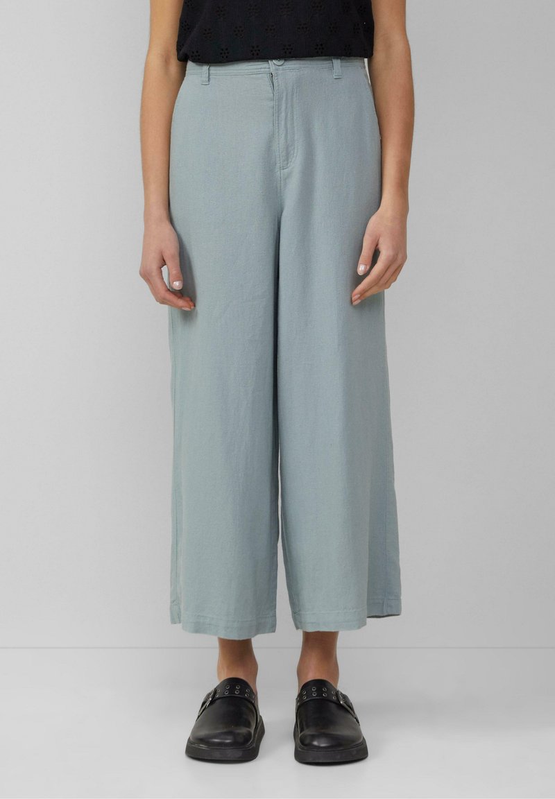 Femme portant un pantalon large court bleu clair, un haut noir sans manches et des chaussures noires sans lacets, debout devant un fond gris uni.