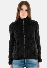 Chaqueta negra con cremallera, de tejido texturizado y suave, con un patrón geométrico y cuello alto, combinada con jeans azules. Apariencia suave y cálida.