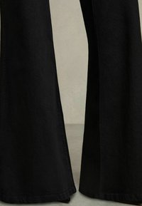 Jean flare noir en denim, avec une surface texturée, des jambes larges et des ourlets bien coupés. Le design inclut un léger détail de couture latérale.