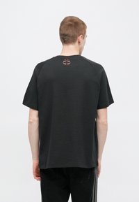 Μαύρο κοντομάνικο μπλουζάκι με στρογγυλή λαιμόκοψη, με ένα μικρό κόκκινο και γκρι λογότυπο στο πίσω μέρος κοντά στη λαιμόκοψη. Κατασκευασμένο από λείο ύφασμα.