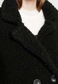 Černý strukturovaný kabát s vykrojeným límcem, velkými knoflíky a hebkým povrchem, přehozený přes bílý jemně žebrovaný svetr.