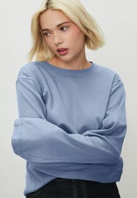Bluza de culoare albastră deschisă, cu guler rotund, umeri lăsați și croială lejeră. Material moale cu detalii de cusătură vizibile.