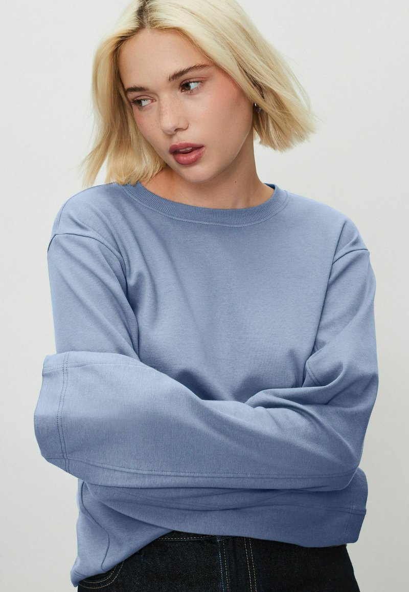 Bluza de culoare albastră deschisă, cu guler rotund, umeri lăsați și croială lejeră. Material moale cu detalii de cusătură vizibile.