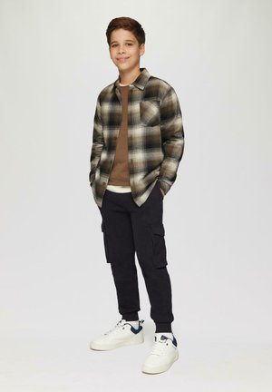 Junge trägt ein kariertes Flanellhemd in Schwarz und Braun über einem braunen Hemd, schwarze Cargo-Hosen und weiße Sneakers mit blauen Akzenten.