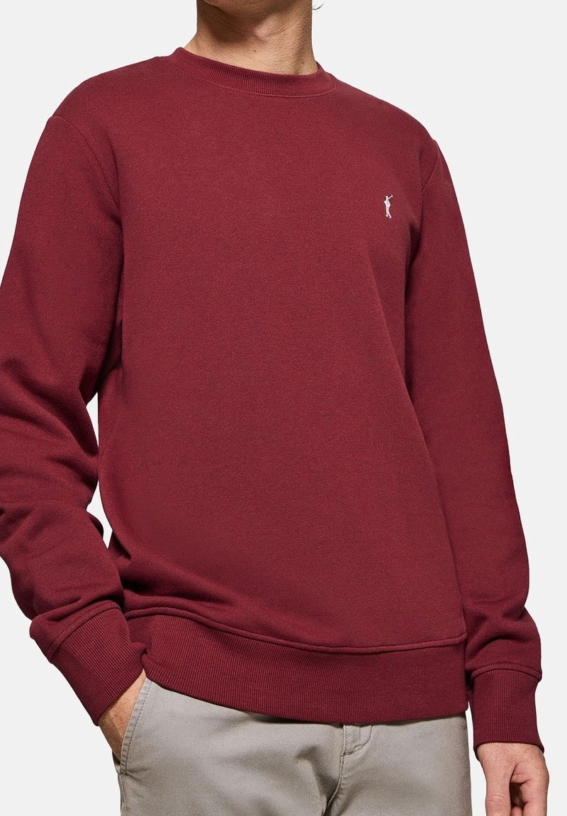 Homme portant un sweat-shirt uni bordeaux à col rond avec poignets et ourlet côtelés, main dans la poche, et petit logo brodé sur la poitrine.