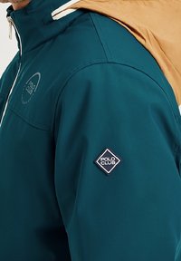 Tealkan färgad jacka med en tanfärgad huva; har en vit dragkedja och en "Polo Club"-logoplack på ärmen. Slätt, lätt material.