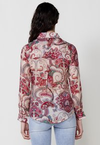 Camicia trasparente a maniche lunghe in tessuto leggero con un motivo paisley rosa e rosso, dotata di colletto con bottoni e una singola tasca sul petto.