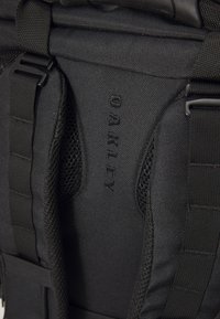 Musta reppu, jossa on kestävä kangas, verkkokohdistuksia, säädettävät olkaimet ja näkyvä kohokuvioitu "OAKLEY"-logo.
