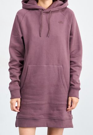 Woman wearing a long mauve hoodie dress with front pocket and drawstring hood, standing against a plain background.