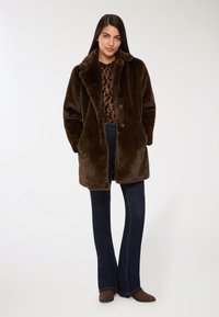 Brązowy płaszcz z sztucznego futra z szerokim kołnierzem, dwoma guzikami i kieszeniami. Połączony z ciemnoniebieskimi dżinsami w stylu dzwonów i brązowymi butami. Widoczny top w zwierzęcy wzór.
