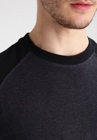 Mörkgrå t-shirt med svarta raglanärmar. Mjuk tygstruktur, klassisk rak halsringning och släta sömmar. Inga mönster eller detaljer synliga.