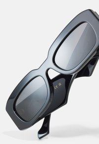 Ochelari de soare negri supradimensionați, cu rame groase din plastic și lentile întunecate. Prezintă o textură netedă și un branding subtil pe partea interioară.