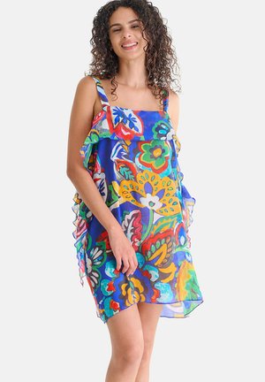 Femme aux cheveux bouclés souriante, portant une robe fleurie colorée sans manches avec un haut à volants, se tenant devant un fond blanc uni.