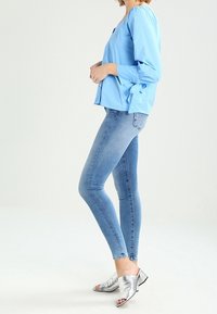 Blusa azzurro chiaro con maniche lunghe, polsini arricciati e dettaglio con lacci, abbinata a jeans skinny azzurro chiaro e mule argento senza tacco.