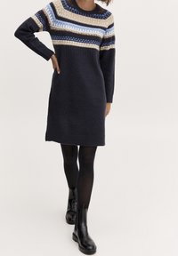 Femme portant une robe-pull bleu marine en maille avec un yoke à motifs bleu, beige et blanc, des collants noirs et des bottines noires, debout avec la main sur la hanche.