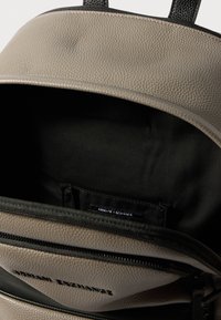 Ανοιχτή γκρι δερμάτινη τσάντα πλάτης με υφή, μαύρη επένδυση εσωτερικά και ετικέτα Armani Exchange στην τσέπη κάτω από το καπάκι.