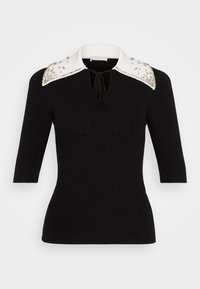 Haut en maille côtelée noire avec un col blanc orné, présentant une encolure en trou de serrure et des manches courtes. Accents décoratifs sur le col.