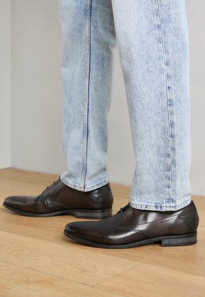 Braune Leder-Hausschuhe mit einem schlanken Design, geschwungener Spitze und dezentem seitlichem Branding. Getragen mit hellblauen Denim-Jeans.