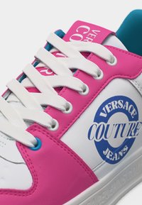 Λευκό αθλητικό παπούτσι με ροζ πάνελ, λευκά κορδόνια, μπλε επένδυση και μπλε λογότυπο "Versace Couture Jeans" στο πλάι.