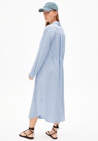 Robe rayée bleu clair avec des manches longues, détails de boutons, une taille nouée et une longueur mi-mollet, associée à des sandales noires à talons et une casquette.