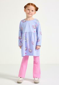 Ung flicka med rött hår i knutar, klädd i en lila klänning med fjärilsmönster över rosa byxor och vita sneakers, står mot en vit bakgrund.