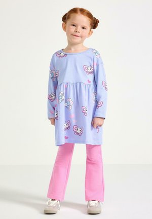 Ung pige med rødt hår sat op i knolde iført en lilla kjole med sommerfugleprint over lyserøde bukser og hvide sneakers, stående mod en hvid baggrund.