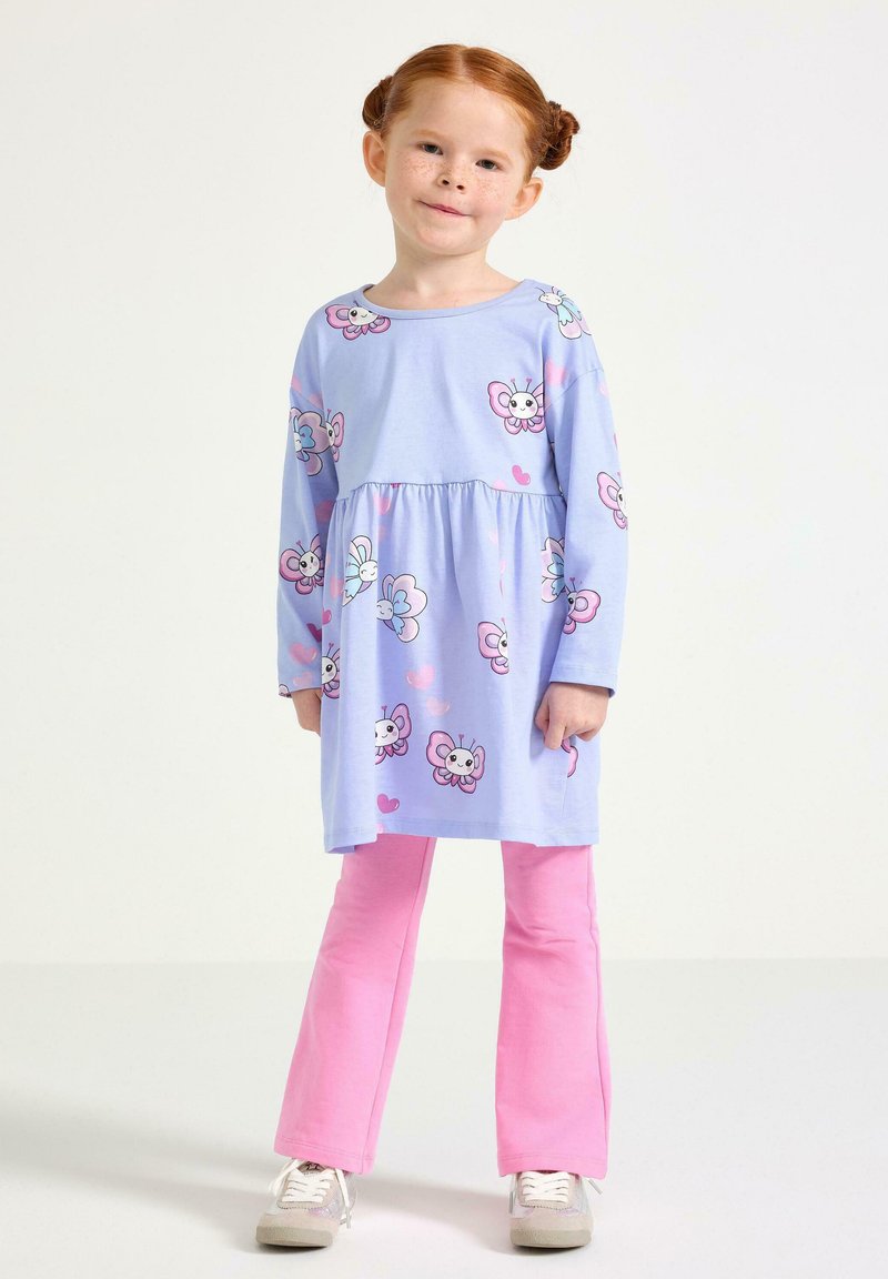 Ung flicka med rött hår i knutar, klädd i en lila klänning med fjärilsmönster över rosa byxor och vita sneakers, står mot en vit bakgrund.